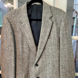 Puede incluir: Una chaqueta de tweed gris con cierre de un solo botón y dos bolsillos con solapa. La chaqueta tiene un patrón de espiga y un forro negro.