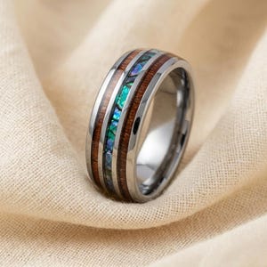 Peut inclure: Une bague en métal argenté poli avec une incrustation centrale de fragments de coquille d'ormeau irisés, entourée de deux bandes de bois brun foncé. La bague présente un profil extérieur lisse et arrondi avec une finition métallique brillante.