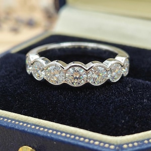 Può includere: Un anello in argento con una fila di pietre preziose rotonde e trasparenti. L'anello è presentato su uno sfondo di velluto blu scuro, all'interno di un cofanetto blu, che ne esalta la brillantezza.