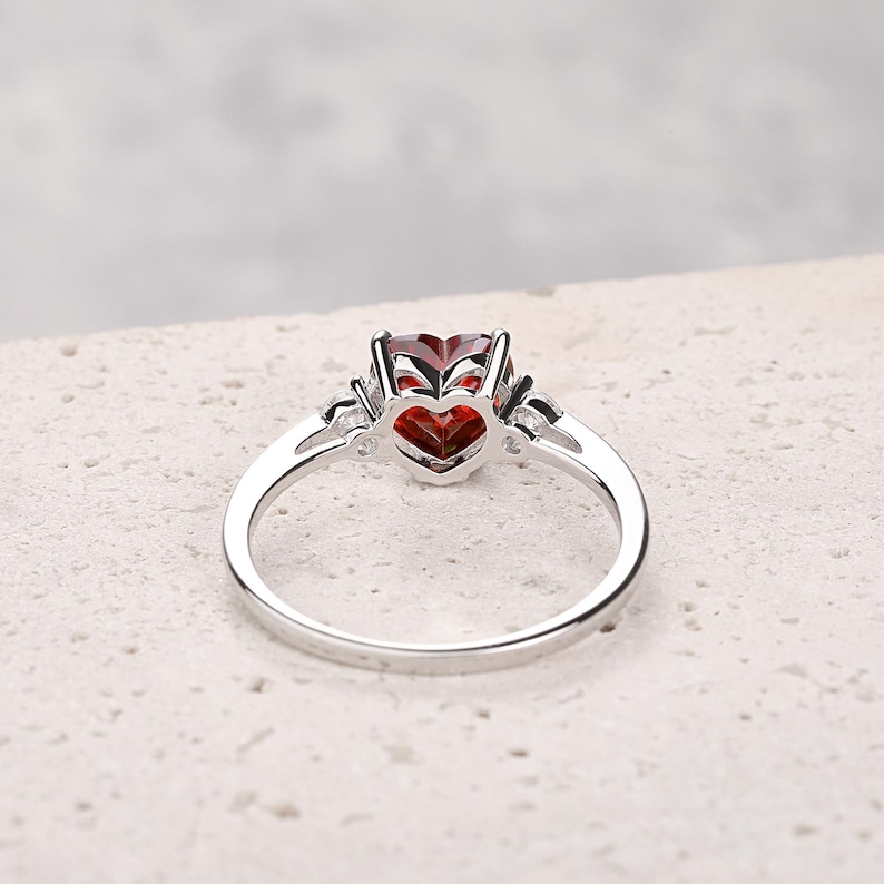 Puede incluir: Un anillo de plata con una piedra preciosa roja en forma de coraz&oacute;n y piedras preciosas m&aacute;s peque&ntilde;as y transparentes en la banda.