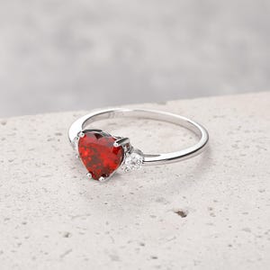 Puede incluir: Un anillo de plata con una gran piedra roja en forma de coraz&oacute;n y dos piedras peque&ntilde;as transparentes en la banda.
