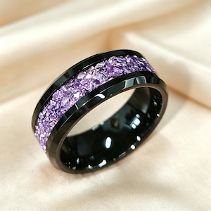 May include: A black ring with a band of crushed purple stones. The ring has a polished black finish and a modern design. The purple stones are set between two black bands, creating a striking contrast. This ring is a piece of jewelry.
