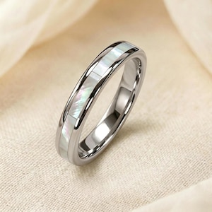 Può includere: Un anello d'argento con una fascia di intarsio in madreperla iridescente. L'anello ha una superficie lucida e riflettente e un design classico ed elegante. L'anello è probabilmente realizzato in acciaio inossidabile o metallo simile.