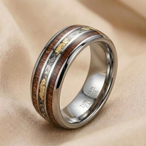 Peut inclure: Une bague en carbure de tungstène argenté avec un incrustation de grain de bois et de feuilles d'or. La bague a une finition polie et les mots "Tungsten Carbide" sont gravés sur le côté. La bague est de style alliance.