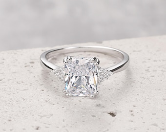 Puede incluir: Un anillo de plata con una gran piedra preciosa rectangular transparente. La piedra preciosa está rodeada de piedras preciosas más pequeñas y transparentes.