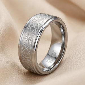 Könnte beinhalten: Ein silberfarbener Ring aus Wolframkarbid mit gebürsteter Oberfläche und einem durchgehenden, eingravierten keltischen Knotenmuster auf dem Außenband. Die Innenseite des Rings ist poliert und trägt die Prägung TUNGSTEN CARBIDE.