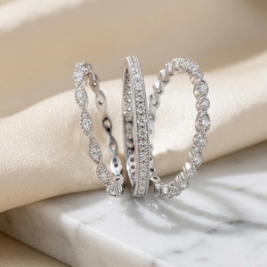 Può includere: Tre anelli color argento con pietre trasparenti. L'anello a sinistra ha un motivo a forma di marquise, quello centrale è una fascia dritta con incastonatura a pavé e quello a destra presenta un design con bordo smerlato e piccole pietre tonde.