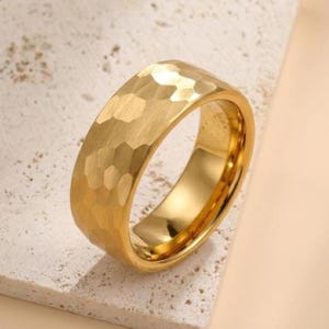 Puede incluir: Un anillo dorado con textura martillada. El anillo tiene una banda ancha y un interior pulido. El anillo se muestra sobre una superficie clara y texturizada. El anillo es una joya.