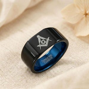 Può includere: Un anello nero in carburo di tungsteno con una fascia interna blu e un'incisione bianca del simbolo massonico della squadra e del compasso con la lettera G. Le parole TUNGSTEN CARBIDE sono chiaramente visibili sulla superficie interna blu.