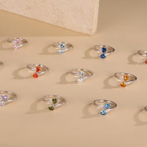 Puede incluir: Una colección de doce anillos de tono plateado, cada uno con dos pequeñas piedras preciosas en forma de corazón de varios colores, incluyendo rosa, azul, naranja, morado, amarillo, verde y transparente, con diseño abierto.