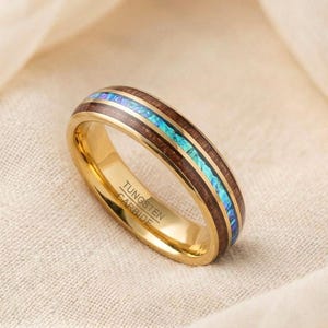 Peut inclure: Une bague en carbure de tungstène de couleur or avec une incrustation de grain de bois et une bande centrale de matériau irisé bleu et turquoise. La bague porte l'inscription "Tungsten Carbide" sur le côté.