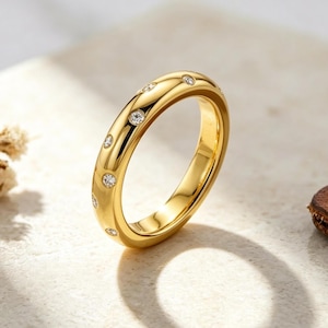 Peut inclure: Une bague dorée ornée de petits diamants clairs, espacés régulièrement. La bague présente une surface lisse et polie, avec un anneau arrondi. Les pierres sont rondes et affleurantes. La bague est présentée sur une surface claire.