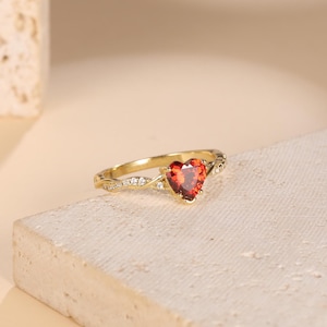 Op de afbeelding: Een gouden ring met een hartvormige rode edelsteen en kleinere heldere edelstenen op de band.
