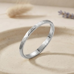 Può includere: Un anello in carburo di tungsteno color argento caratterizzato da una texture martellata sulla superficie esterna. L'interno della fascia è liscio e chiaramente inciso con il testo TUNGSTEN CARBIDE, che indica la composizione del metallo.