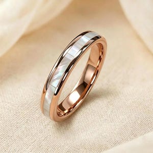 May include: Rose gold ring with a band of white mother-of-pearl inlay. The ring is set against a soft, neutral background, highlighting the polished metal and iridescent sheen of the inlay. A simple, elegant design.