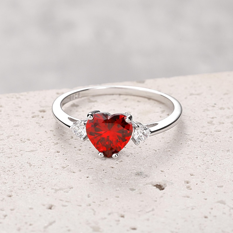 Puede incluir: Un anillo de plata con una gran piedra preciosa roja en forma de coraz&oacute;n y dos piedras preciosas transparentes m&aacute;s peque&ntilde;as a cada lado.