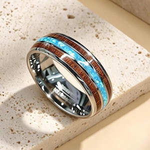 May include: A silver ring with a wood grain and blue inlay design. The ring features a polished silver band with a central inlay of light blue material, framed by bands of dark brown wood grain. The ring is displayed on a textured surface.