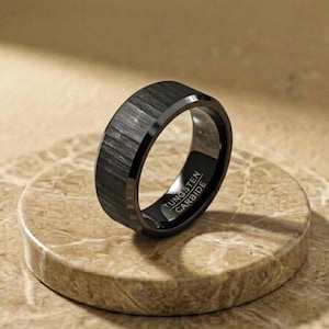 May include: A black ring with a textured, brushed finish on the outer surface. The interior of the band is smooth and polished, featuring the laser-engraved text TUNGSTEN CARBIDE. The ring has a flat profile with beveled edges on the outer band.