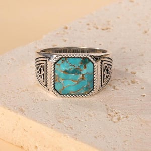 Può includere: Un anello d'argento con una grande pietra turchese rettangolare. L'anello presenta un design dettagliato con motivi a nodo celtico sui lati e un bordo a forma di corda attorno alla pietra. Il testo "IN THE PALM OF HIS HAND" è inciso sull'anello.