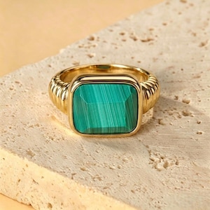 Può includere: Un anello d'oro con una pietra rettangolare verde smeraldo. L'anello ha una fascia strutturata e un'incastonatura quadrata attorno alla pietra. La pietra presenta un motivo a strisce. L'anello è posto su una superficie chiara e strutturata.