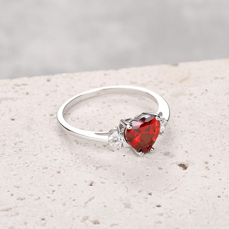 Puede incluir: Un anillo de plata con una gran piedra preciosa roja en forma de coraz&oacute;n y dos piedras preciosas m&aacute;s peque&ntilde;as transparentes en la banda.