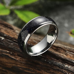 May include: A silver-colored ring with a dark blue, speckled inlay. The ring is set against a natural wood background, highlighting its polished metal and the textured surface of the inlay. The ring's design is simple yet elegant.