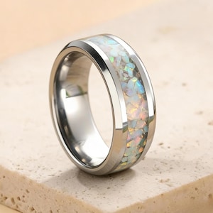 Può includere: Un anello color argento con una fascia larga. Il centro dell'anello è riempito di pietre iridescenti, bianche e dai colori pastello. L'anello ha una finitura lucida e riflettente e le pietre sembrano essere a filo con il metallo.