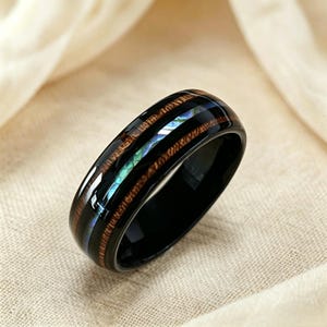 Può includere: Un anello nero con finitura lucida. L'anello presenta una fascia in legno e un intarsio di conchiglia di abalone iridescente. Il legno ha un motivo a venature naturali. L'anello è posto su uno sfondo di tessuto neutro.