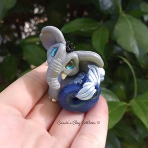 Puede incluir: Una pequeña figurita de elefante gris con ojos azules y una cola azul y plateada con purpurina. La figurita está sentada en una mano.