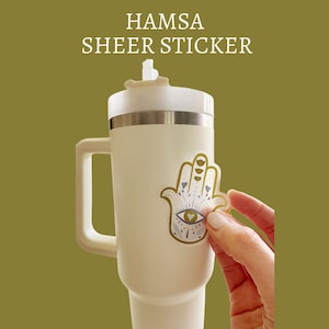 May include: A white travel mug with a gold Hamsa hand sticker with a blue eye and hearts. The sticker is being applied to the mug.