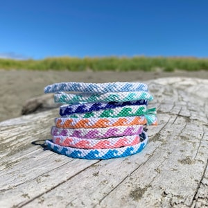 May include: A stack of colourful friendship bracelets with a wave pattern, made with embroidery floss. The bracelets are arranged on a weathered piece of driftwood.