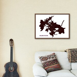 May include: A framed map of College Station, Texas, with a brown frame and a white background. The map is in maroon and shows the city's streets and landmarks. The map is hanging on a beige wall above a white couch with a patterned pillow.