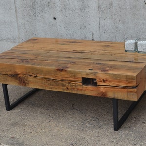 Pu&ograve; includere: Un tavolino da caff&egrave; in legno rustico con un piano in legno riciclato e gambe in metallo nero. Il tavolino &egrave; su una superficie in cemento.