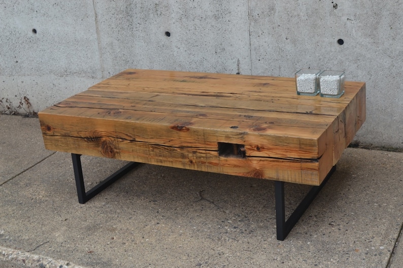 Pu&ograve; includere: Un tavolino da caff&egrave; in legno rustico con un piano in legno riciclato e gambe in metallo nero. Il tavolino &egrave; su una superficie in cemento.