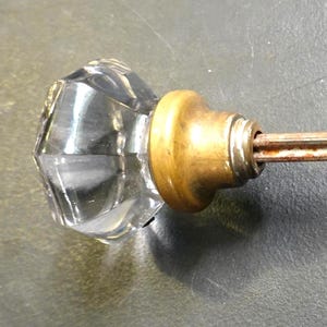 May include: A clear glass door knob with a faceted design. The knob is attached to a brass base and a metal rod. The door knob is a vintage style.