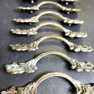 May include: A collection of antique bronze-toned drawer pulls. Each pull features an arched handle and ornate, floral-inspired end pieces. The hardware is arranged in rows, showcasing their aged patina and decorative design.
