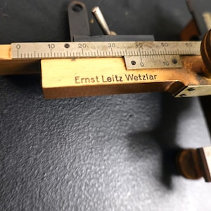 May include: A vintage wooden and brass measuring tool with a metal ruler marked in increments. The tool has the text "Ernst Leitz Wetzlar" engraved on the wooden body. The tool is used for precise measurements.