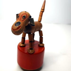 May include: A vintage wooden dog toy with a red base. The dog is brown with painted facial features, including large eyes and a smiling mouth. The tail is segmented and upright. The toy is a classic example of mid-century design.