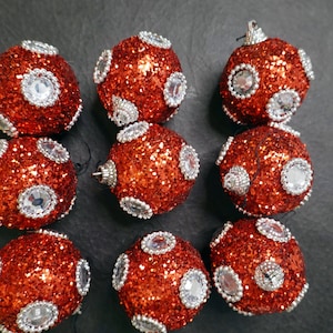 Pode incluir: Oito enfeites de Natal vermelhos com glitter prateado e strass transparentes. Os enfeites estão dispostos em duas filas de quatro.