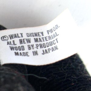 K&ouml;nnte beinhalten: Nahaufnahme eines wei&szlig;en Etiketts mit schwarzem Text. Das Etikett lautet "&copy; WALT DISNEY PROD. ALL NEW MATERIAL WOOD BY-PRODUCT MADE IN JAPAN". Das Etikett ist an einem schwarzen, strukturierten Stoff befestigt.
