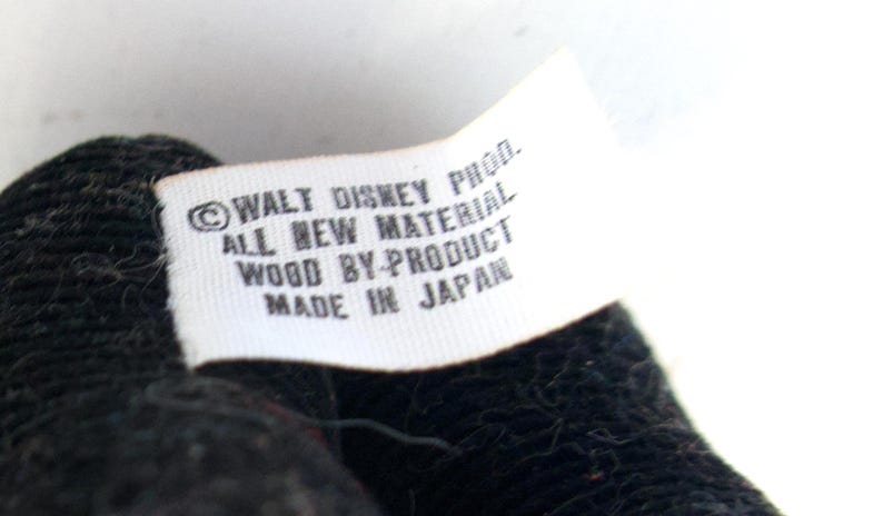 K&ouml;nnte beinhalten: Nahaufnahme eines wei&szlig;en Etiketts mit schwarzem Text. Das Etikett lautet "&copy; WALT DISNEY PROD. ALL NEW MATERIAL WOOD BY-PRODUCT MADE IN JAPAN". Das Etikett ist an einem schwarzen, strukturierten Stoff befestigt.