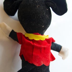 K&ouml;nnte beinhalten: Eine Vintage-Minnie-Maus-Puppe, von hinten gesehen. Die Puppe hat einen schwarzen K&ouml;rper, gro&szlig;e Ohren und ein rotes Kleid mit gelbem Kragen. Die Arme der Puppe sind schwarz mit wei&szlig;en Handschuhen. Eine gelbe Schleife sitzt oben auf dem Kopf.