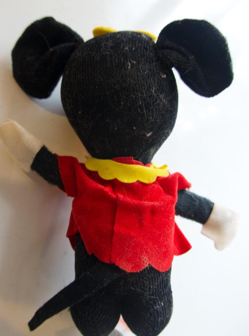 K&ouml;nnte beinhalten: Eine Vintage-Minnie-Maus-Puppe, von hinten gesehen. Die Puppe hat einen schwarzen K&ouml;rper, gro&szlig;e Ohren und ein rotes Kleid mit gelbem Kragen. Die Arme der Puppe sind schwarz mit wei&szlig;en Handschuhen. Eine gelbe Schleife sitzt oben auf dem Kopf.