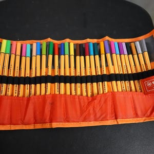 May include: An orange pen case containing a selection of colourful Stabilo pens. The pens are arranged in rows, with colours such as green, blue, pink, and purple. The case has a black strap and a red Stabilo label.