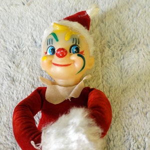Puede incluir: Un muñeco de elfo navideño vintage con un gorro rojo y blanco, un atuendo rojo y blanco y un ribete de piel blanca. El elfo tiene una cara pintada con detalles verdes, amarillos y azules.