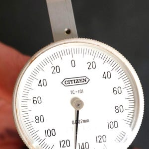 May include: A Citizen TC-101 dial indicator with a white face and black markings. The dial reads from 0 to 140, with increments marked in white. The indicator measures in 0.002mm increments. The device has a silver metal arm and a black base.