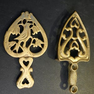 May include: Two antique brass trivets. One is heart-shaped with a bird and floral design. The other is iron-shaped with heart and swirl cutouts. Both have handles for easy use. Decorative kitchenware.