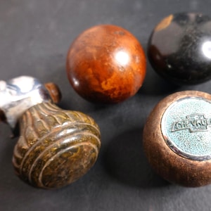 May include: Four vintage car knobs. One is a brown and silver gear shift knob. Two are round wooden knobs, one brown and one black. The fourth knob is brown with a Chevrolet emblem.