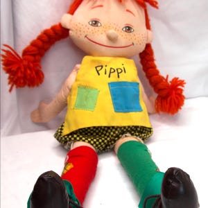 May include: A Pippi Longstocking doll with red yarn hair, freckles, and a smiling face. She wears a yellow dress with the name "Pippi" and two patches, black and white checkered shorts, and mismatched socks and shoes.