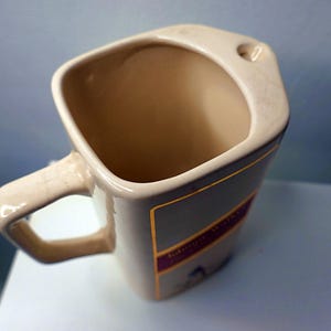 May include: Tan ceramic mug with a square shape and a handle. The mug has a label with gold and purple accents. The interior is a light tan color. The mug is sitting on a white surface.